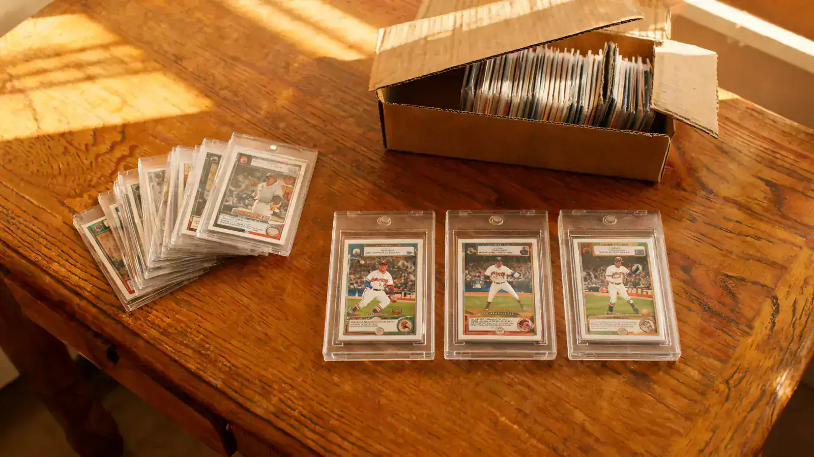 A collector's desk under warm lamp light, rookie cards in Card Saver 1 holders and Ultra Pro one-touches, BCW storage box visible at the edge