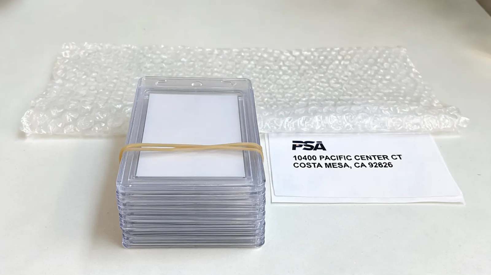 A bundle of twenty Card Saver 1 holders secured with a rubber band, placed next to a printed PSA submission label and bubble wrap