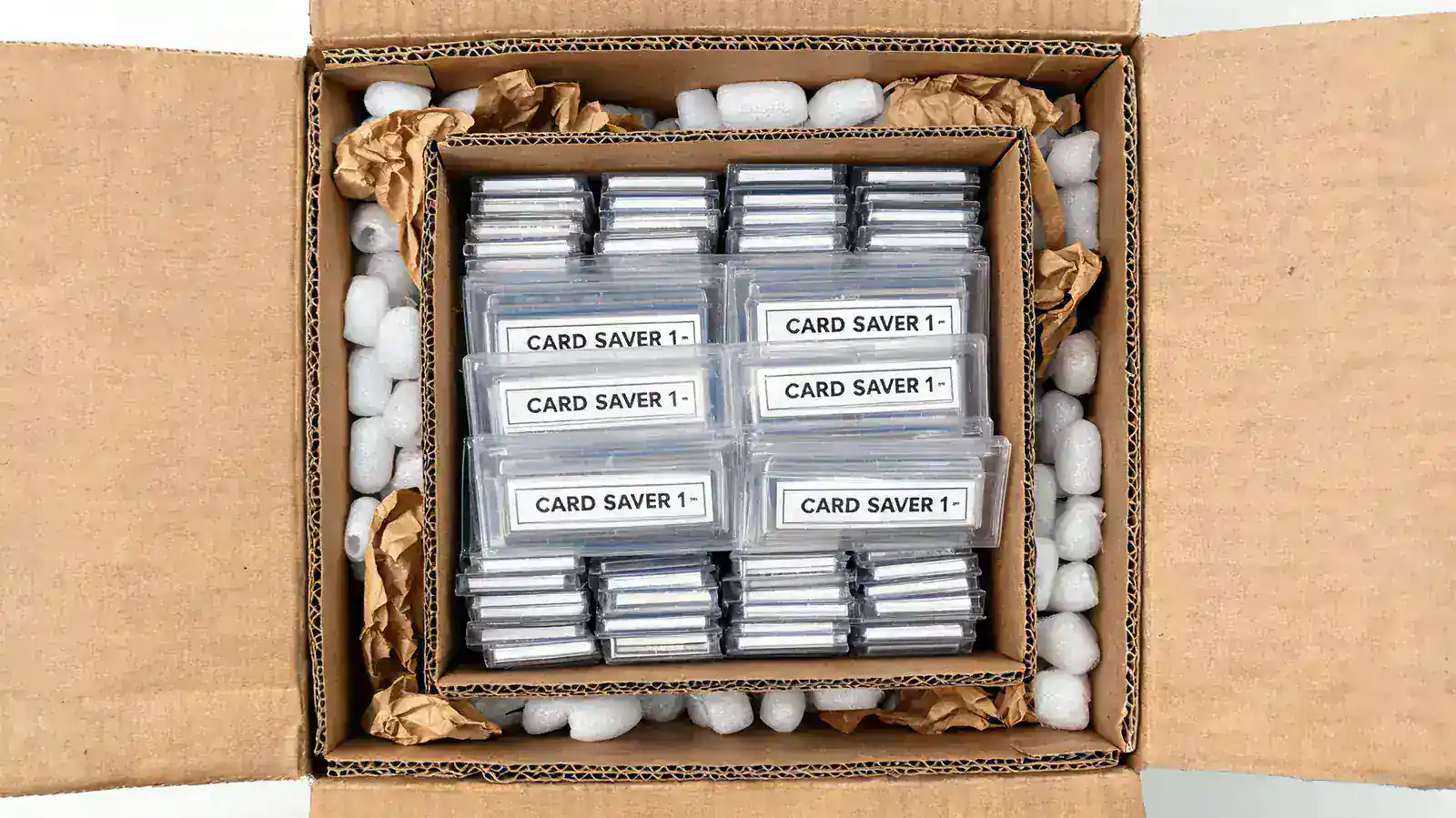 A small inner box packed tight with card bundles sitting inside a larger outer shipping box filled with packing peanuts, demonstrating the box-within-a-box shipping method